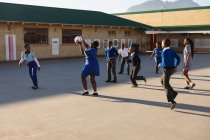 Вид сбоку на группу африканских школьниц и школьников, прыгающих и играющих с мячом на детской площадке в детской школе — стоковое фото