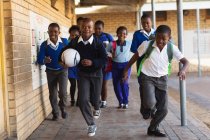 Вид спереди на группу молодых африканских школьников, бегущих во дворе школы со школьными сумками и футболом на игрушечном корабле — стоковое фото