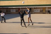 Vista trasera de una joven colegiala africana y dos colegiales saltando y jugando con una pelota en el patio de recreo en una escuela primaria del municipio - foto de stock