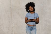 Vista frontale di una giovane donna di razza mista appoggiata a un muro usando uno smartphone e indossando cuffie, ascoltando musica all'aperto al sole — Foto stock