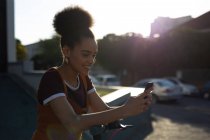 Vista lateral de cerca de una joven mujer de raza mixta sonriente sentada en una pared en la calle usando un teléfono inteligente, retroiluminado por la luz del sol - foto de stock