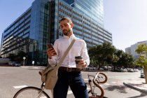 Vue de face rapprochée d'un jeune homme caucasien tenant un café à emporter et utilisant un smartphone, appuyé sur son vélo dans une rue de la ville. Nomade numérique en mouvement . — Photo de stock