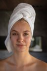 Portrait close up of a young Caucasian woman wearing a towel on her hair, looking straight to camera in a modern bathroom. — Stock Photo