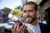 Vue de face gros plan d'un jeune homme caucasien souriant parlant sur un smartphone le tenant devant son visage et portant des écouteurs dans une rue de la ville. Nomade numérique en mouvement . — Photo de stock
