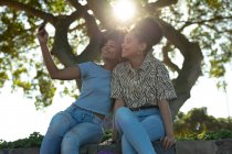 Vista frontal close-up de duas jovens sorridentes irmãs de raça mista adultas sentadas em uma parede em um parque urbano, usando um smartphone e tirando selfies, retroiluminadas com lente de sinalização — Fotografia de Stock