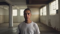 Vue de face d'un jeune hispano-américain percé d'une chemise blanche ordinaire regardant attentivement la caméra dans un entrepôt vide — Photo de stock