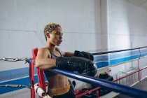 Side view of female boxer resting in the corner of the boxing ring at boxing club. Strong female fighter in boxing gym training hard. — Stock Photo