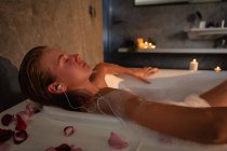 Vista laterale ravvicinata di una giovane donna caucasica distesa nella vasca da bagno con gli occhi chiusi ad ascoltare musica con gli auricolari . — Foto stock