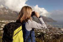 Side view close up of a mature Caucasian woman wearing a backpack standing and looking at the view through a pair of binoculars during a hike. — Stock Photo
