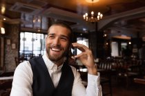 Vista frontal de cerca de un joven caucásico sonriente en una llamada telefónica sentado en una mesa dentro de un café. Nómada digital en movimiento . - foto de stock