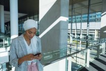 Empresária feliz em hijab apoiando-se em trilhos e usando telefone celular no corredor no escritório moderno — Fotografia de Stock