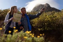 Side view close up of a mature Caucasian woman and man, the woman holding a map and the man pointing to the distance during a hike in the countryside, a mountain peak and cloud in the background — Stock Photo
