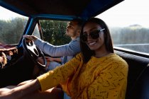 Close up side view of a young mixed race couple sitting in their pick-up truck during a road trip. The man is driving and the woman is turning to camera and smiling — Stock Photo