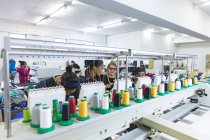 Вид спереду на різноманітну групу жінок-колег, які працюють за рядом автоматизованих швейних машин на заводі спортивного одягу . — стокове фото
