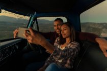 Vista frontale di una giovane coppia mista sorridente seduta in macchina a farsi un selfie al crepuscolo, durante una sosta durante un viaggio su strada. L'interno dell'auto è illuminato con luci ad arco . — Foto stock