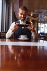 Vue de face gros plan d'un jeune homme caucasien souriant utilisant son smartphone assis à une table avec une tasse de café à l'intérieur d'un café. Nomade numérique en mouvement . — Photo de stock