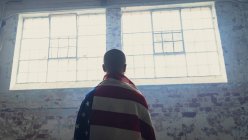 Rear view of a young man with back turned against the camera and an American flag over shoulders inside an empty warehouse — Stock Photo