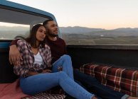Vista laterale di una giovane coppia mista seduta fuori nel retro del pick-up, che abbraccia e gode della vista al tramonto durante una sosta durante un viaggio su strada . — Foto stock