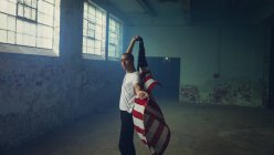 Vista lateral de um jovem hispânico-americano vestindo uma camisa branca simples segurando uma bandeira americana dentro de um armazém vazio — Fotografia de Stock