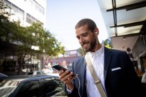 Vue de face rapprochée d'un jeune homme caucasien souriant utilisant un smartphone et portant des écouteurs dans une rue de la ville. Nomade numérique en mouvement . — Photo de stock
