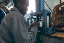 Seitenansicht eines jungen afrikanisch-amerikanischen Arbeiters, der in einer Fabrik sitzt und eine Maschine bedient, die Cricketbälle herstellt, im Hintergrund ist ein Kollege zu sehen, der neben ihm am Fließband arbeitet.. — Stockfoto