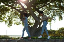 Nahaufnahme von zwei jungen lächelnden erwachsenen Mischlingsschwestern, die mit ihren unter ihnen geparkten Elektrorollern an einer Mauer in einem Stadtpark entlang laufen — Stockfoto