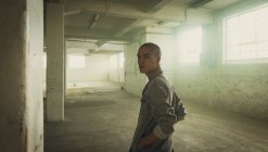 Vue latérale d'un jeune hispano-américain percé d'une veste grise avec les mains dans les poches, regardant attentivement la caméra à l'intérieur d'un entrepôt vide — Photo de stock