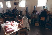 Erhöhte Seitenansicht eines jungen afrikanisch-amerikanischen Mannes mit Hut, der an einer Werkbank sitzt und mit ausgeschnittenen roten Lederformen in einer Werkstatt einer Cricketfabrik arbeitet, im Hintergrund arbeiten Kollegen — Stockfoto