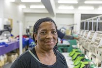 Retrato close-up de uma mulher de meia-idade de raça mista em pé ao lado de uma fileira de máquinas em uma fábrica de roupas esportivas brilhantemente iluminada, olhando para a câmera e sorrindo com uma linha de produção no fundo . — Fotografia de Stock
