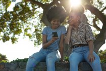 Vista frontal close-up de duas jovens irmãs de raça mista adultas sorridentes sentadas em uma parede em um parque urbano, usando um smartphone e compartilhando fones de ouvido para ouvir música, retroiluminadas com brilho de lente — Fotografia de Stock
