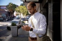 Vista lateral de perto de um jovem caucasiano vestindo uma bolsa de ombro usando um smartphone, em pé no pavimento em uma rua da cidade. Digital Nomad em movimento . — Fotografia de Stock