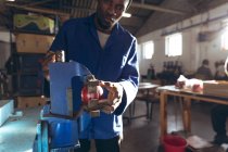Вид спереди молодой афроамериканец, работающий на заводе, делающий крикетные мячи, держащий крикетный мяч в тисках и смотрящий в камеру . — стоковое фото