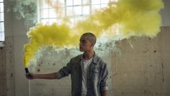 Frontansicht eines jungen hispanisch-amerikanischen Mannes, der eine graue Jacke über einem weißen Hemd trägt und von der Kamera wegschaut, während er eine Rauchmaschine in der Hand hält, die gelben Rauch in einer leeren Lagerhalle produziert — Stockfoto
