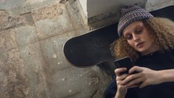 Hochwinkelaufnahme einer jungen kaukasischen Frau mit lockigem Haar, die ein schwarzes Hemd und eine Mütze trägt und mit dem Kopf auf einem Skateboard auf dem Boden liegt, während sie ein Mobiltelefon in einer leeren Lagerhalle benutzt — Stockfoto