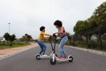 Vista laterale di due giovani sorelle di razza mista che cavalcano su scooter elettrici su una strada tranquilla — Foto stock