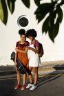 Vista frontal de dos hermanas adultas jóvenes de raza mixta, una llevando una mochila y la otra sosteniendo un monopatín, sonriendo y mirando a un teléfono inteligente, de pie en una calle al sol - foto de stock