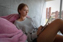 Seitenansicht einer jungen kaukasischen Frau, die mit einem Tablet-Computer auf einem Sofa in einer Wohnung liegt. sie entspannt sich und praktiziert Selbstpflege. — Stockfoto
