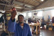 Vue de face gros plan d'un jeune Afro-Américain tenant une balle pour la vérifier dans un atelier dans une usine de fabrication de balles de cricket, en arrière-plan collègues travaillent à un établi sur d'autres parties de la chaîne de production
. — Photo de stock