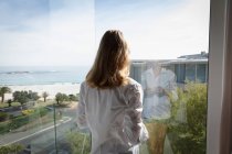 Назад думку крупним планом молода кавказька жінка носить білу сорочку, що стоїть біля вікна тримає чашку кави і дивлячись, море і пляж у фоновому режимі. — стокове фото