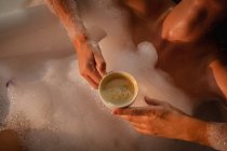 Close up mid section of woman holding a cup of coffee and lying back in a foam bath. — Stock Photo