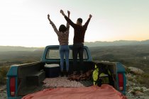 Vue arrière d'un jeune couple mixte debout à l'arrière de sa camionnette, les bras levés, profitant de la vue au coucher du soleil lors d'un arrêt sur la route . — Photo de stock