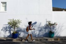 Vue latérale d'une jeune femme métisse portant un sac à dos parlant sur un smartphone qu'elle tient devant elle, tout en marchant dans une rue urbaine au soleil — Photo de stock
