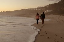 Side view of a mature Caucasian man and woman holding hands and walking together along a beach beside the sea at sunset — Stock Photo