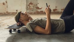 Vista lateral da vista lateral de um jovem hispânico-americano com piercings vestindo uma camisa cinza escura e gorro deitado no chão com a cabeça em um skate e usando um telefone celular — Fotografia de Stock