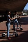 Vista laterale di due giovani sorelle di razza mista adulte in piedi con scooter elettrici in un parco urbano, guardando la fotocamera e sorridendo — Foto stock