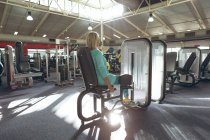 Вид сзади на активную пожилую женщину-инвалида, занимающуюся с тренажером для завивки ног в фитнес-студии — стоковое фото