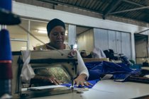 Vue de face gros plan d'une femme de race mixte d'âge moyen assise et conduisant une machine à coudre dans une usine de vêtements de sport . — Photo de stock