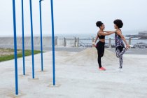 Вид збоку на двох молодих сестер-перегонів у спортивному одязі, що стоять, спираючись один на одного і розтягуючи ноги у зовнішньому міському просторі — стокове фото