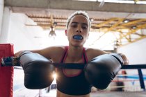 Close-up de boxeador feminino com protetor bucal olhando para a câmera enquanto estava no ringue de boxe. Forte lutador feminino no treinamento de ginásio de boxe duro . — Fotografia de Stock