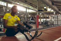 Vista frontal de un atlético afroamericano discapacitado sentado en la pista de carreras en el gimnasio - foto de stock